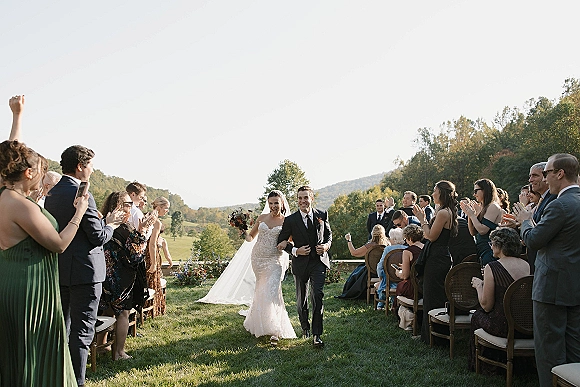 Wedding recessional as bride and groom walk hand in hand down the aisle, bouquet and veil flowing, guests clapping on a mountain lawn backdrop