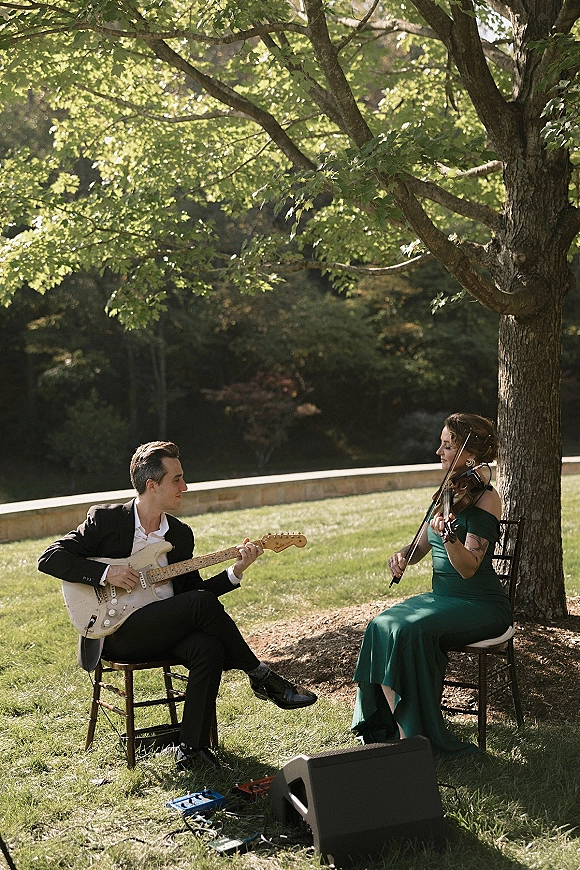 Wedding musicians, a violinist in a green dress and guitarist in a suit, perform under a tree on a lawn with sound equipment and cables