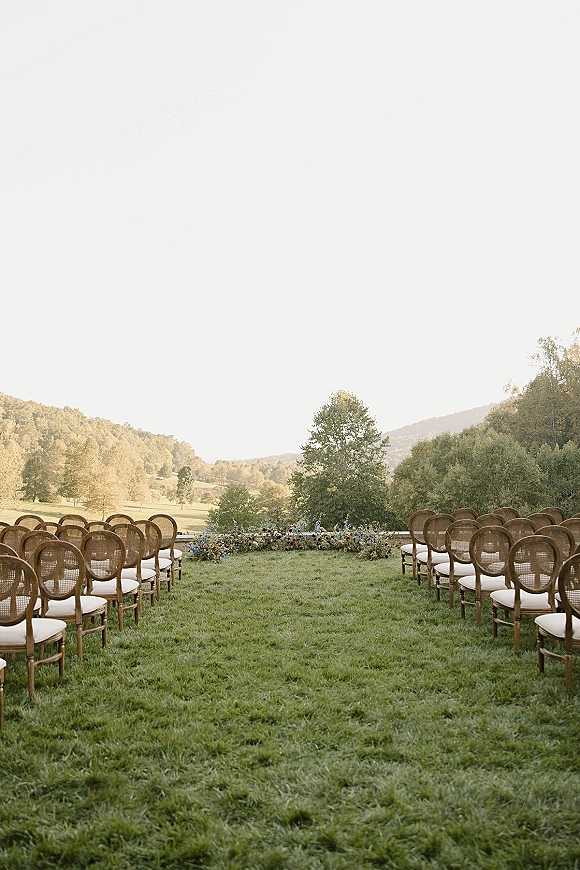 Outdoor ceremony setup with wedding ceremony seating in rows of wood and cane-back chairs, a greenery ground floral arrangement, and valley hills beyond