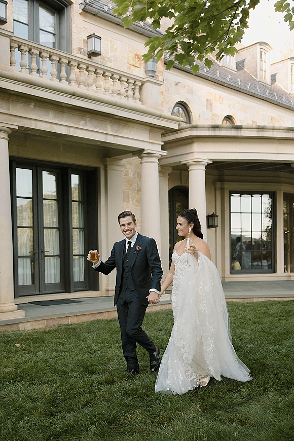 Wedding couple portrait of bride and groom walking hand in hand, toasting with champagne and whiskey outside a stone estate with columns