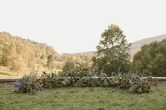 Ceremony floral arrangement with grounded altar flowers in a wildflower meadow style along a stone wall, set on a lawn with rolling hills beyond