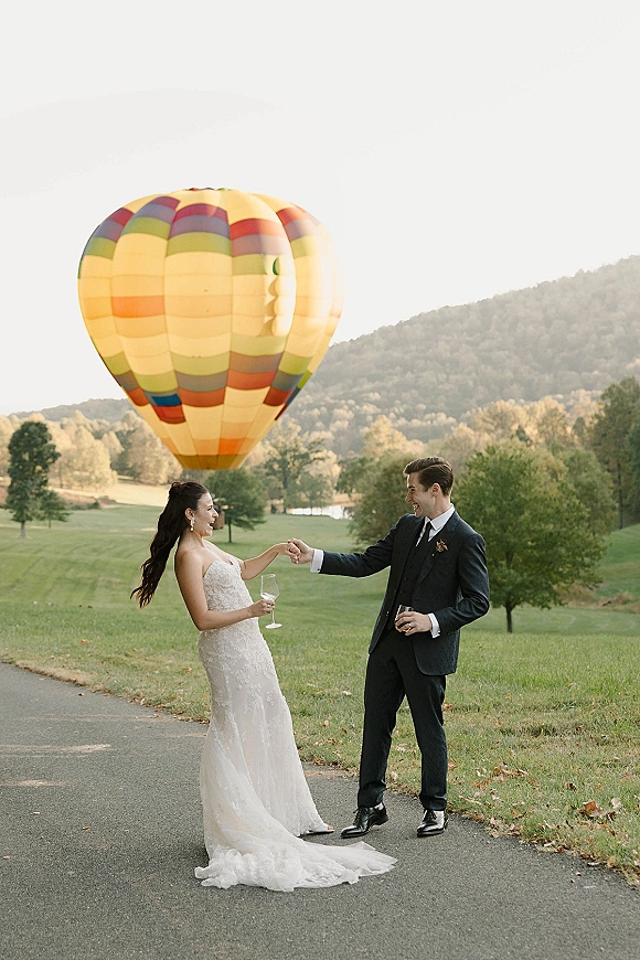 Couple portrait of bride and groom toasting with champagne in a grassy field, lace dress and boutonniere, hot air balloon behind them
