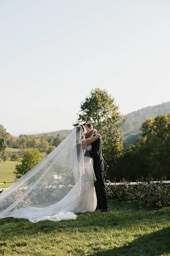 Wedding kiss portrait of bride and groom kissing as her cathedral veil blows in the wind on a meadow with mountains and rolling hills behind