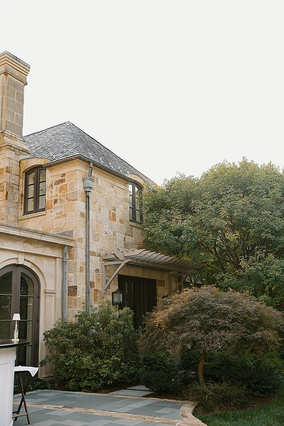 Wedding venue exterior with a cocktail table draped in white, set on a stone patio beside a manor facade with arched windows and pergola awning