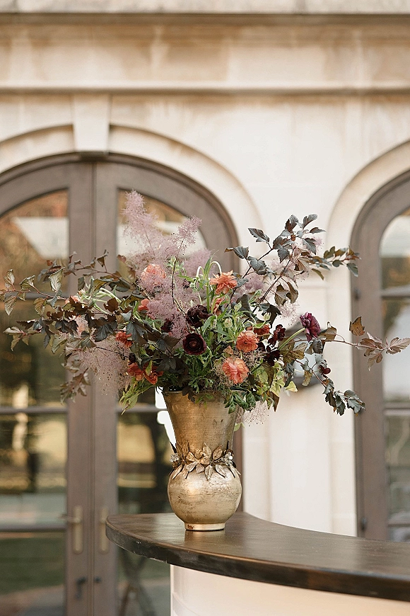 Wedding floral arrangement statement floral centerpiece with branchy greenery in a gold vase on a bar counter before arched doors and windows