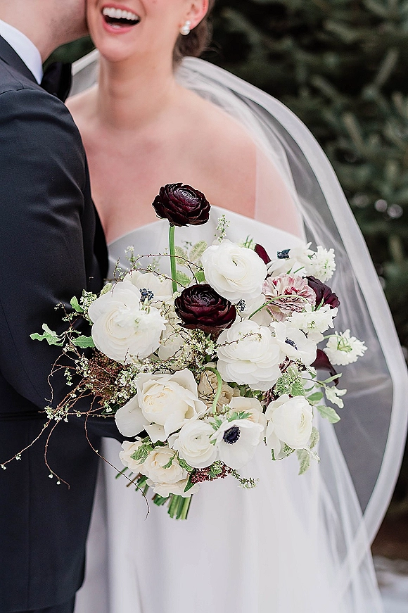 Bridal bouquet of burgundy and white blooms with greenery, held by a bride in veil and strapless dress beside groom in suit outdoors