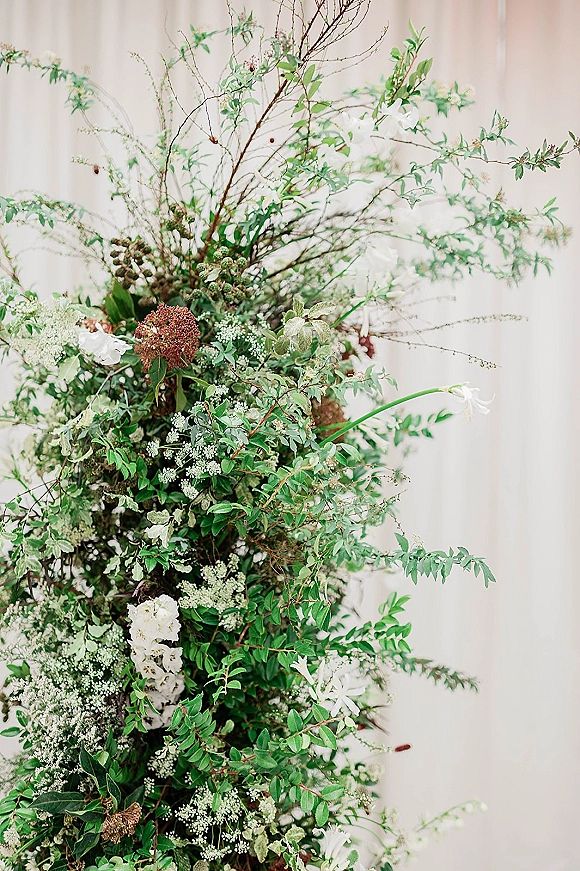 Wedding floral arrangement with greenery, branches, and airy white flowers against a white curtain backdrop for a ceremony installation