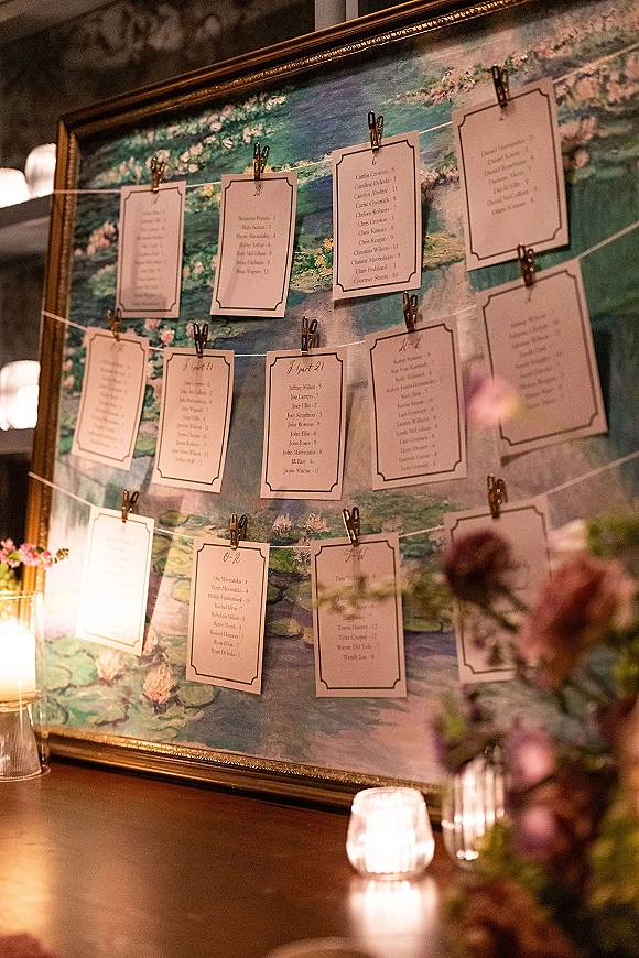 Wedding seating chart escort card display in a gold frame with mini clothespins on string, candles and florals on a wooden table before a painted canvas backdrop