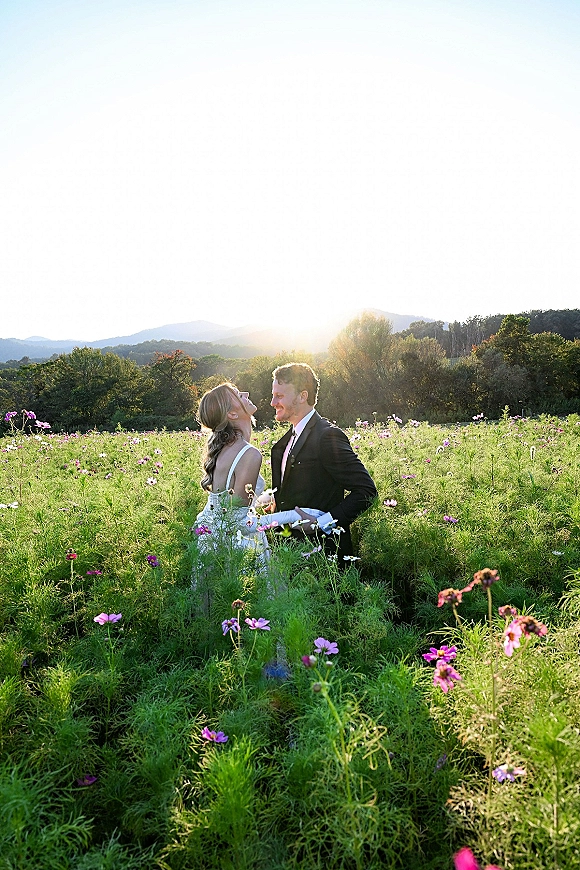 Couple portrait of bride and groom outdoors, bride in strapless gown holding bouquet, smiling in tall grass with mountains at sunset
