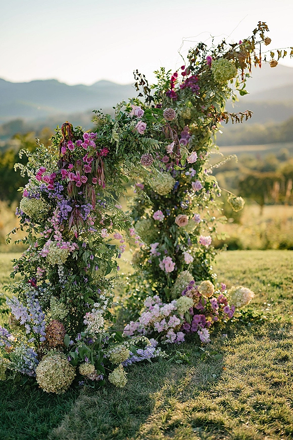 Wedding ceremony arch with asymmetrical floral arch of roses and hydrangea, set on a grassy lawn with mountains in golden hour light