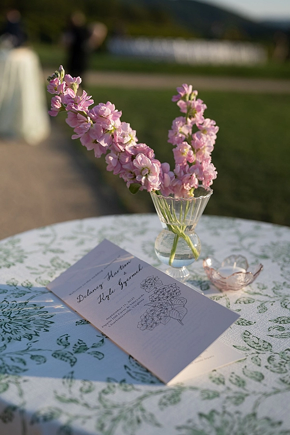 Invitation suite wedding invitation flat lay on patterned tablecloth with pink bud vase and tea light, lawn ceremony chairs and hills behind