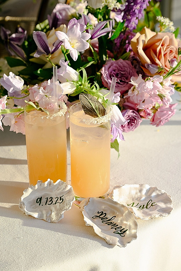 Signature wedding cocktails beside his and hers cocktails in highball glasses with salted rims, sage leaves, and straws on a sunlit table with roses