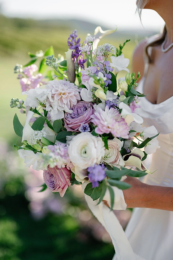 Bridal bouquet of pastel wedding bouquet blooms with lavender and white flowers, greenery, and trailing ribbon held against a blurred outdoor field