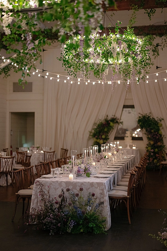 Reception tablescape with a long banquet table setup, taper candles, glassware, and hanging greenery beneath string lights indoors