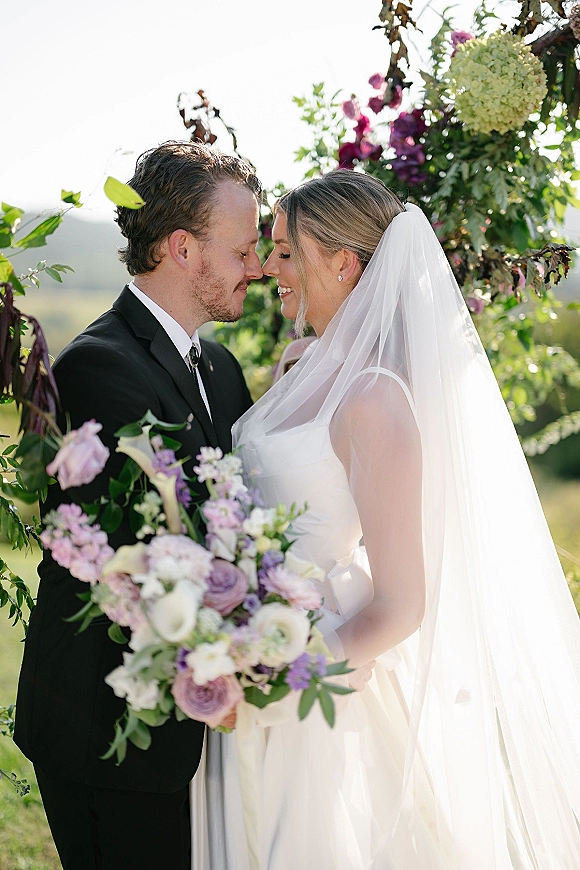 Couple portrait of bride and groom close up with foreheads touching, bride in veil holding calla lily bouquet beneath floral arch and hills
