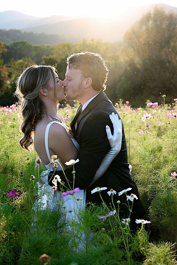 Wedding kiss portrait of newlyweds kissing in a wildflower meadow at sunset, bride in white opera gloves and groom in black tuxedo with mountains behind