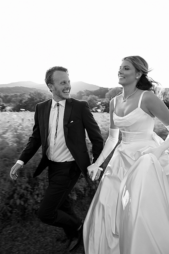 Couple portrait in a black and white wedding portrait, bride in satin dress and gloves holding hands with groom in bolo tie in a mountain field