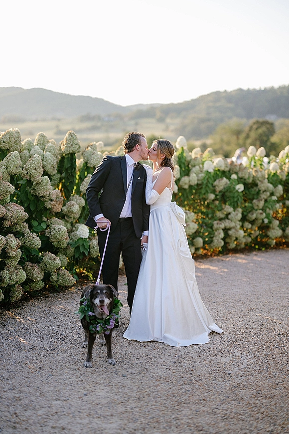 Wedding couple portrait of bride and groom kissing in bridal gloves and black suit, holding a leashed dog with floral collar on a mountain path