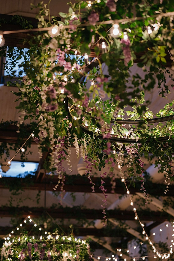 Wedding ceiling decor with hanging floral installation, greenery garlands, string lights, and chandelier rings under rustic wood beams