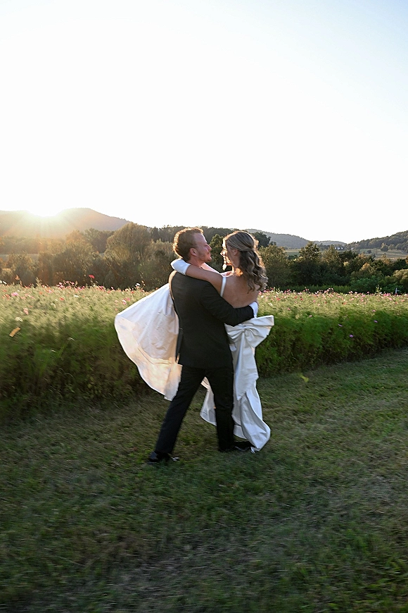 Wedding couple portrait of groom carrying bride in a strapless dress with long train and veil in the wind at sunset meadow with wildflowers