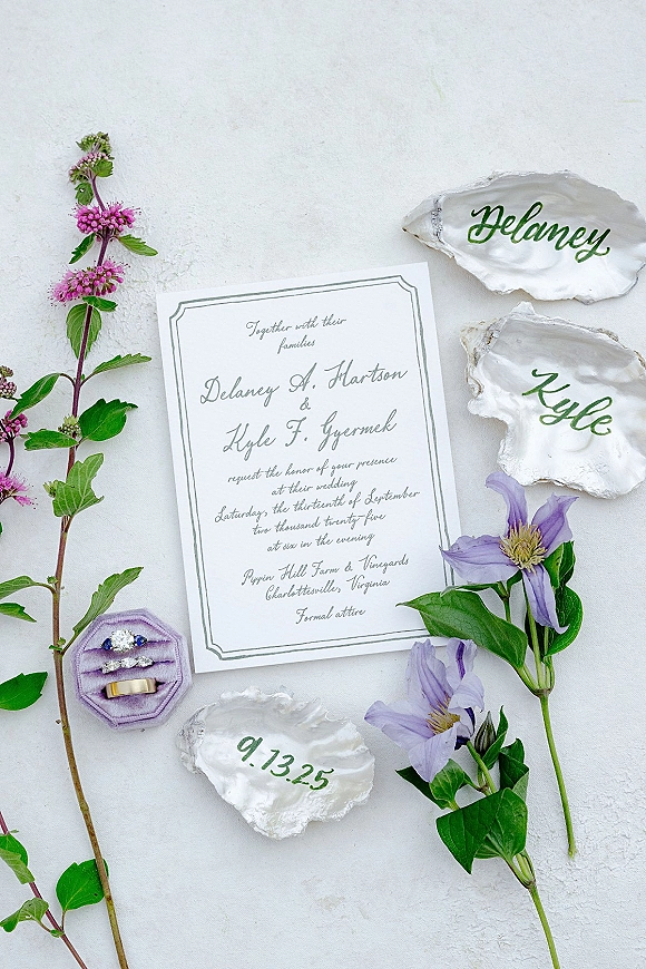 Wedding invitation suite with calligraphy flat lay, oyster shell place cards and rings in a lavender box, styled with purple and pink flowers on white texture