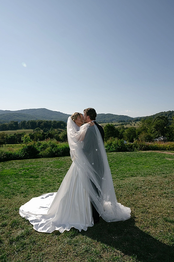 Wedding kiss as the bride and groom kissing under a wind-blown veil, her long train trailing in a grassy meadow with mountains beyond