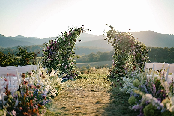 Ceremony altar with a floral ceremony arch of greenery and purple-pink blooms, lined aisle florals and white chairs on a lawn with mountain views