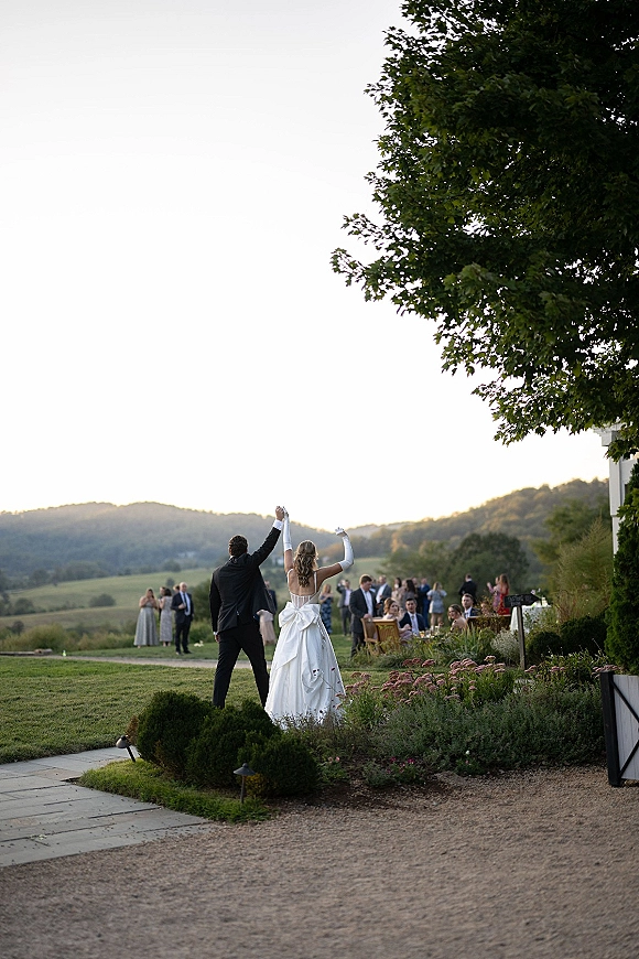 Newlywed exit as bride and groom walk away with hands raised, bride in strapless satin gown and opera gloves on a garden path