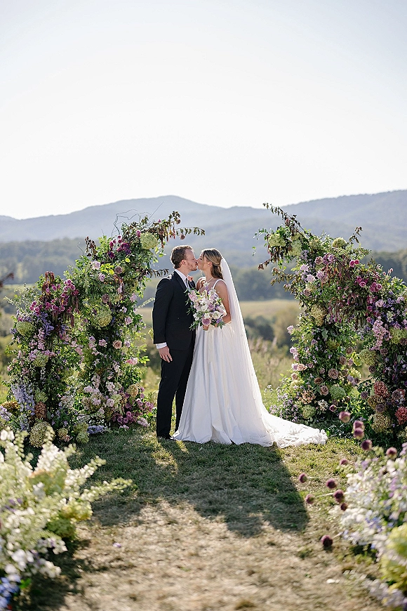 Wedding kiss at an outdoor ceremony kiss under a hydrangea and rose floral arch, mountain landscape behind, bride in veil holding bouquet