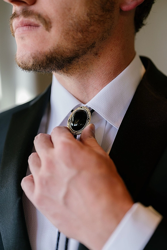 Groom getting ready, adjusting tie with a bolo tie clasp over a white dress shirt and black suit jacket in soft window light indoors