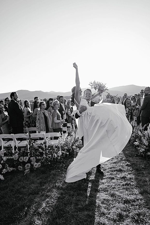 Recessional moment as groom carries bride down an outdoor aisle, her bouquet raised, veil streaming, guests cheering with mountains behind