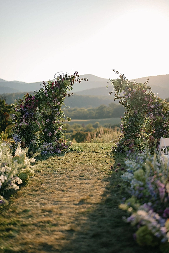 Ceremony aisle decor with wildflower arrangements and greenery framing floral pillars, lined by white chairs on a lawn with mountain views
