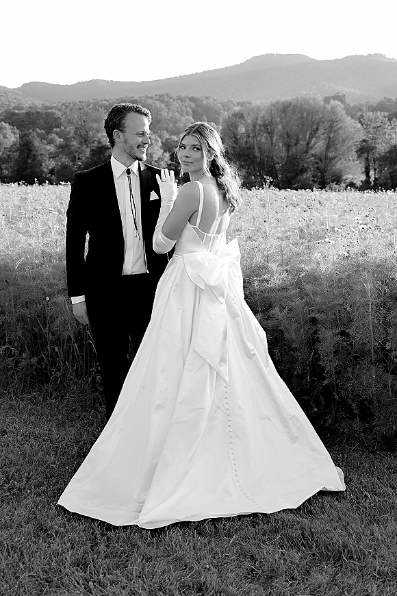 Couple portrait in a black and white wedding portrait style, bride in bow-back gown and gloves holding groom’s lapel in a mountain meadow
