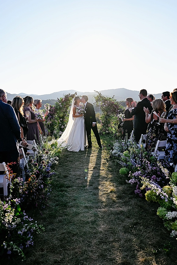 Ceremony kiss as bride in strapless gown and long veil kisses groom in black tux under floral arch with mountain landscape behind