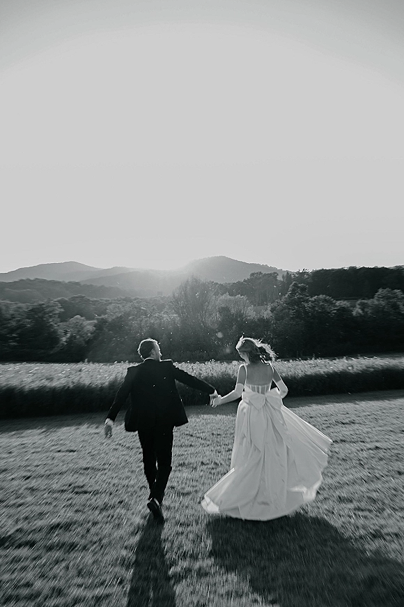 Couple portrait of bride and groom walking away holding hands, her wedding dress train and veil flowing in sunset-lit mountain meadow