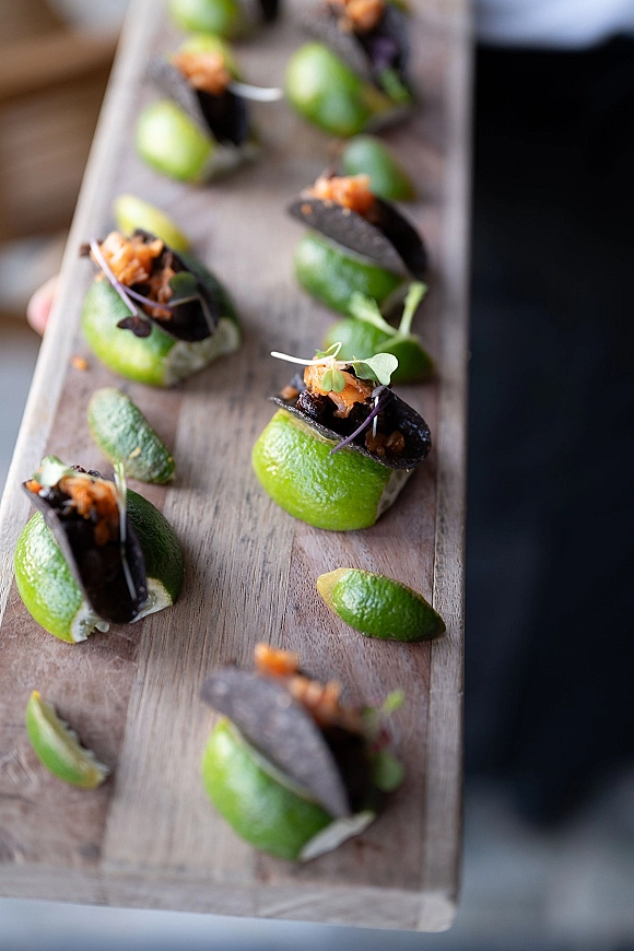 Wedding appetizers of bite-size black taco shells with microgreens and lime on a wood serving board, hand-held tray in blurred indoor setting