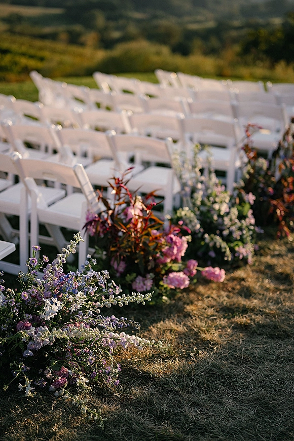 Ceremony aisle decor with white folding chairs and pink and purple meadow florals lining the aisle on a grassy hillside field