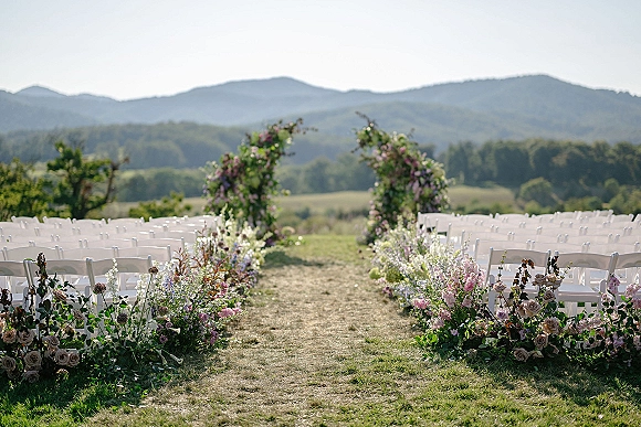 Ceremony setup for an outdoor wedding ceremony with a floral arch, white folding chairs, and aisle flowers on a grassy field with mountains beyond