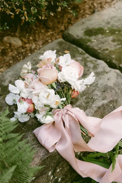 Bridal bouquet of blush and white blooms with greenery and berry accents, wrapped in a satin ribbon, resting on stone steps in a garden setting