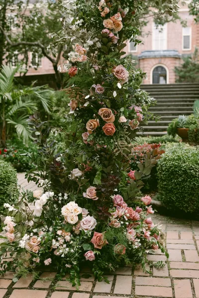 Wedding floral arch with blush and peach roses and lush greenery, set along a brick walkway in a garden courtyard by stone steps