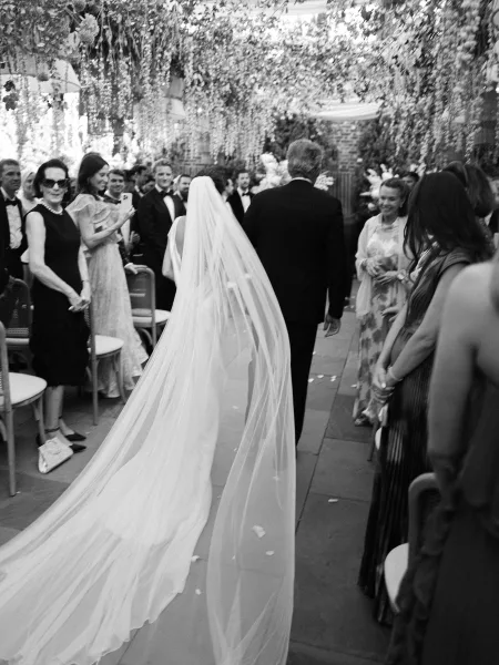Wedding processional with a bride escorted down the aisle along a petal-strewn stone floor, as seated guests watch against a brick wall.