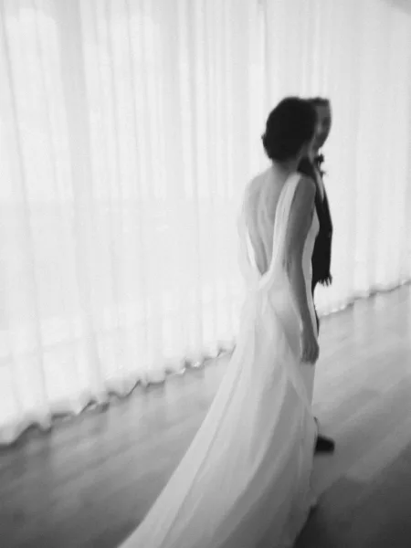Couple portrait of a walking bride and groom, her dress train trailing as they cross a hardwood floor before sheer curtains.
