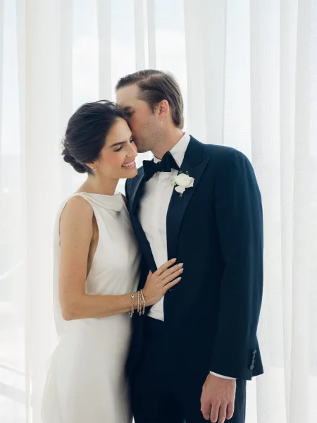 Couple portrait of a groom kissing the bride on the forehead as they embrace, his black bow tie catching soft light by a sheer curtained window.
