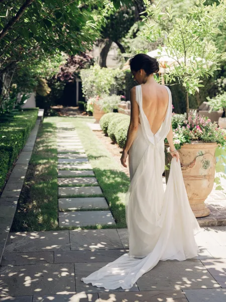 Bridal portrait of a bride walking away in an open back wedding dress, gently lifting her dress train along a stone walkway in a garden.