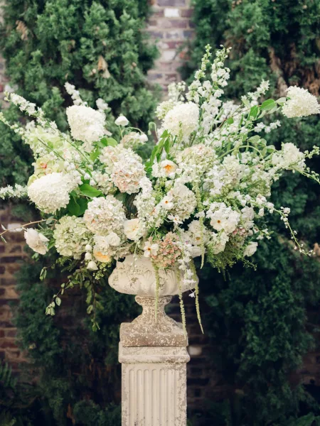 Wedding floral arrangement spilling from a classic urn, clustered with lush blooms, set against a brick wall and flanked by evergreen trees.