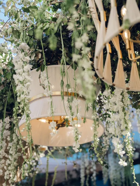 Hanging floral installation with a floral chandelier design, accented by brass hardware, suspended beneath wooden beams against a blue sky.