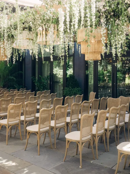 Ceremony setup highlighting a thoughtful ceremony seating arrangement with cane-back chairs on a stone patio beside glass doors and potted plants.