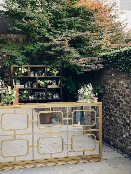 Wedding bar setup for an outdoor cocktail bar featuring a mirrored bar, set against ivy-draped brick with garden trees and a stone patio.