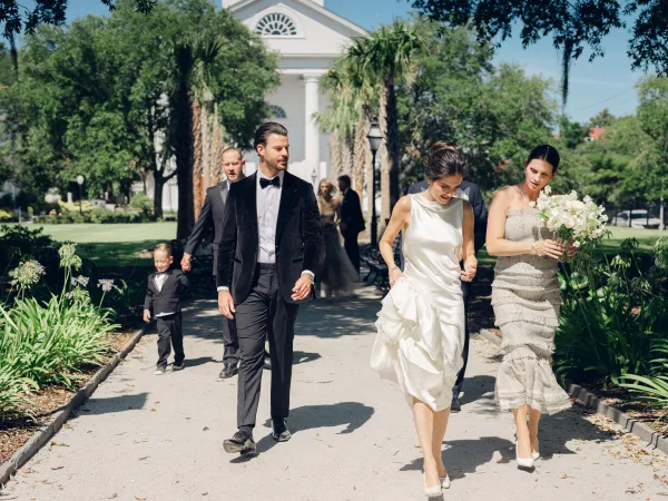 Wedding party portrait with bride and groom walking along a garden path by a white church and palm trees under a blue sky, bride carrying a bouquet.