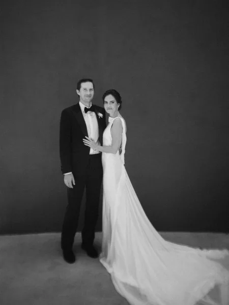 Couple portrait in a studio wedding portrait setting, bride with a sleek bridal silhouette and groom in black tie with a bow tie, on a dark backdrop.